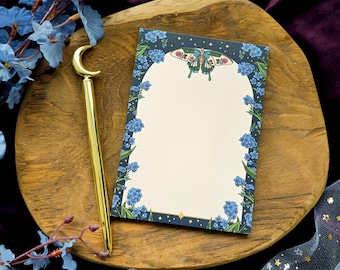 Puede incluir: Un bolígrafo dorado con una parte superior en forma de media luna descansa junto a un bloc de notas. El bloc de notas presenta un borde floral de flores azules y una ilustración de mariposa. Los artículos están sobre una bandeja de madera, con flores azules y una tela transparente con estrellas doradas en el fondo.