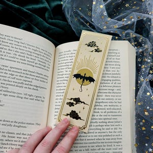 Può includere: Un segnalibro decorativo con bordo dorato presenta una silhouette di drago contro un sole, con nuvole. Il segnalibro è posto in un libro aperto, con uno sfondo verde scuro e un tessuto trasparente con stelle dorate.