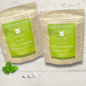 Puede incluir: Dos bolsas de papel marrón con etiquetas verdes que contienen tabletas de pasta dental natural. Las etiquetas muestran un hipopótamo de dibujos animados y el texto "Happi Hippo plant organics Peppermint". Las bolsas están sobre una superficie de madera blanca con una ramita de hojas de menta verde y tres tabletas blancas.