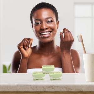 May include: A woman smiling while flossing her teeth. Three boxes of vegan dental floss are in front of her. A toothbrush is in a white cup to the right. The boxes have green lids and the text "Vegan Care Dental Floss" on them.