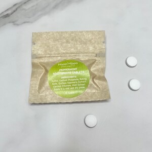 Könnte beinhalten: Ein kleiner brauner Papierbeutel mit einem grün-weißen Etikett, auf dem "Happi Hippo plant organics PEPPERMINT TOOTHPASTE TABLETS" steht und die Zutaten aufgelistet sind. Neben dem Beutel liegen drei weiße runde Zahnpasta-Tabletten.
