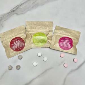 Może przedstawiać: Trzy małe brązowe papierowe torebki z białymi etykietami zawierające naturalne tabletki do pasty do zębów. Etykiety mają nazwę marki "Happi Hippo" i wymieniają smaki: cynamon, mięta pieprzowa i truskawka. Na białej powierzchni rozrzucone są również białe i różowe okrągłe tabletki.