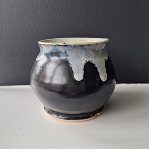 May include: A black ceramic mug with a white glaze drip design. The mug has a rounded body and a slightly flared rim.