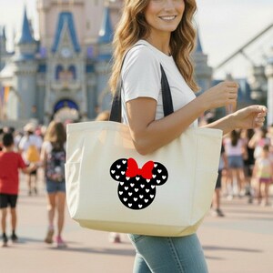 May include: A cream-colored tote bag with black straps featuring a Minnie Mouse design. The design includes a black silhouette of Minnie's head filled with white hearts and a red bow. The bag is carried by a person in front of a castle.
