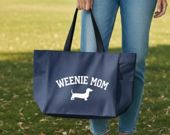 May include: Navy blue tote bag with black handles and side pockets. The bag features the text "WEENIE MOM" above a white dachshund silhouette. The bag is held in front of a person wearing blue jeans and white sneakers.