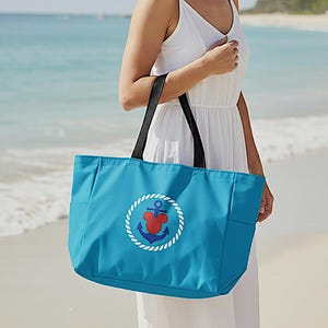 May include: A turquoise tote bag with black handles and a nautical anchor design. The anchor is red and blue, surrounded by a white rope circle. The bag has side pockets and is held by a person on a beach.