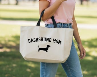 May include: A beige tote bag with black handles, featuring the text "DACHSHUND MOM" above a black silhouette of a dachshund. The bag is carried by a person wearing a pink shirt and blue jeans.