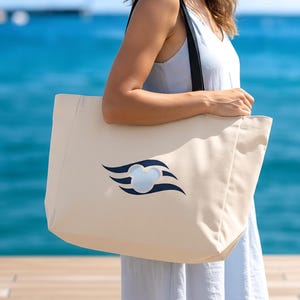 May include: A beige canvas tote bag with a navy blue logo featuring a stylized Mickey Mouse head and curved lines. The bag has black straps and is held in front of a blue ocean background.