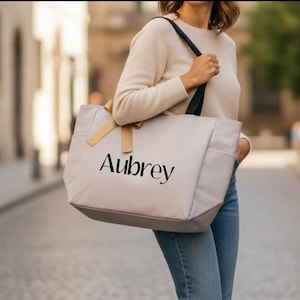 May include: A light gray tote bag with black straps and the name "Aubrey" printed in black. The bag has a light brown bow and side pockets. The person is wearing a cream-colored sweater and blue jeans.