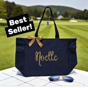 May include: A navy blue tote bag featuring the name Noelle in gold script lettering, a gold ribbon bow attached to the top left, and black shoulder straps. A black sign with the white text Best Seller is visible, and sunglasses are below.
