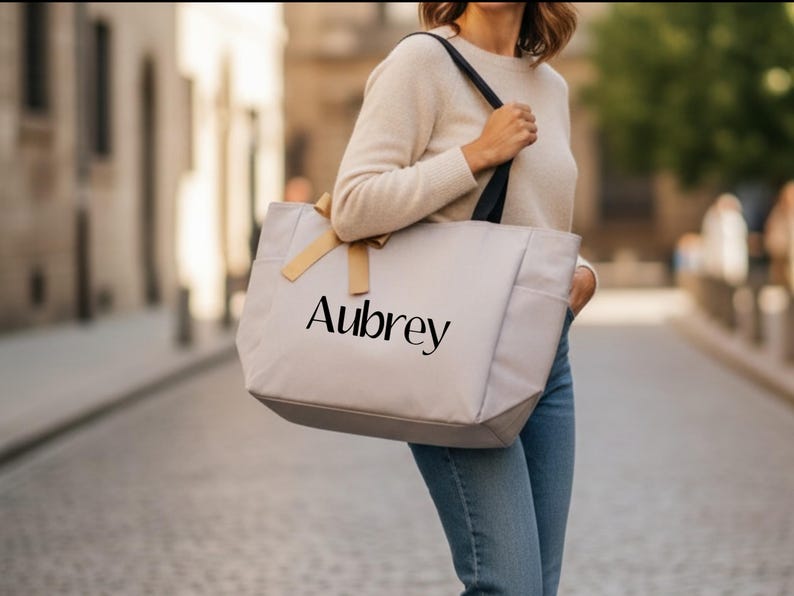 May include: A light gray tote bag with black straps and the name "Aubrey" printed in black. The bag has a light brown bow and side pockets. The person is wearing a cream-colored sweater and blue jeans.
