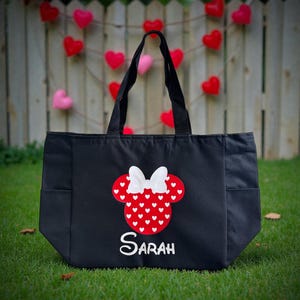 May include: Black tote bag with a Minnie Mouse design. The design features a red head shape with white heart accents and a white bow. The name "Sarah" is written in white script below the design. The bag has two side pockets and black handles.