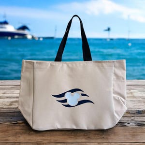 May include: Beige tote bag with black handles and a side pocket. The bag features a navy blue and light blue Mickey Mouse logo. The background shows a blue ocean and a boat.