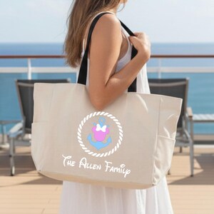 May include: A beige tote bag with black straps features a nautical design with an anchor, bow, and rope circle. The text "The Allen Family" is printed below the design. The bag has side pockets and is held by a person on a deck with a blue ocean background.