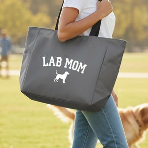 May include: A gray tote bag with the words "LAB MOM" in white block letters and a white silhouette of a Labrador Retriever. The bag has black straps and side pockets. A golden retriever dog is in the background.