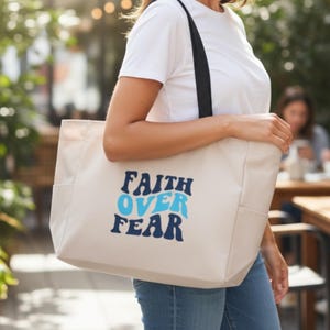May include: A beige tote bag with black straps and side pockets. The bag features the text "FAITH OVER FEAR" in blue and navy blue lettering. The bag is being held by a person wearing a white t-shirt and blue jeans.