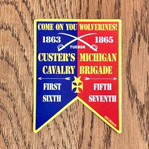 Puede incluir: Un banner amarillo, azul y rojo con el texto "COME ON YOU WOLVERINES!"  "1863" "TUEBOR" "1865" "CUSTER'S MICHIGAN CAVALRY BRIGADE" "FIRST SIXTH" "FIFTH SEVENTH" y "1863.DESIGNS".