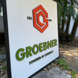 May include: A white sign with a black frame, featuring a red and white logo with the text "EST. 1976". The sign also has the text "GROEBNER POWERED BY EXPERIENCE" in green raised letters.