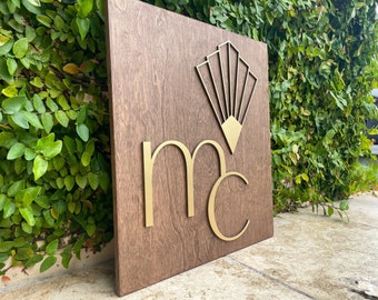 May include: A wooden sign with the letters "MC" in gold, with a stylized gold fan design above the letters.