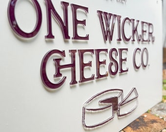 May include: White sign with the text "ONE WICKER CHEESE CO." in raised burgundy letters. The text "Est. 2024" is in smaller white letters above the main text. A stylized image of a wheel of cheese is below the text.