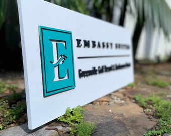 May include: White sign with a teal blue square featuring a stylized letter E with a wave design. The text "Embassy Suites" is printed in black above the square.