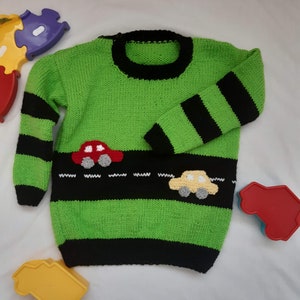 Puede incluir: Suéter de punto verde con rayas negras y un diseño de coche. El suéter tiene un cuello y puños negros.