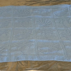 May include: A light blue knitted baby blanket with a repeating pattern of various designs. The blanket is made of soft yarn and has a delicate texture.