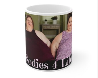 Peut inclure: Une tasse en céramique blanche avec une photo de deux femmes assises sur un canapé. Les femmes portent des vêtements décontractés et sourient. Le texte "Sodies 4 Life" est imprimé au bas de la tasse en rose.
