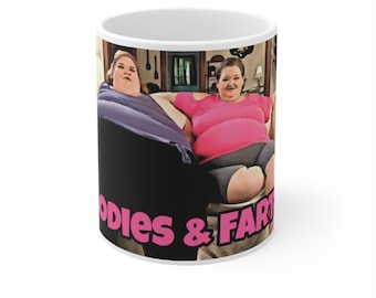 Peut inclure: Une tasse en céramique blanche avec un fond noir représentant deux femmes assises sur un canapé. Les femmes portent des vêtements colorés. Le texte "Bodies & Farts" est imprimé en rose au bas de la tasse.