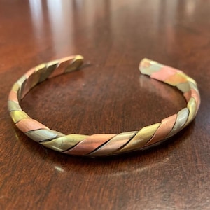 Könnte beinhalten: Ein verdrehtes Kupfer-, Silber- und Goldarmband. Das Armband besteht aus drei verschiedenen Metallen, die miteinander verwoben sind, um ein einzigartiges und stilvolles Design zu schaffen.