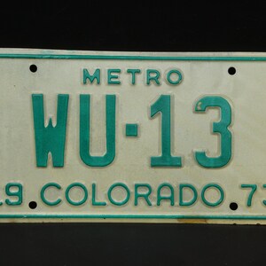 Puede incluir: Una placa de matrícula vintage de Colorado, blanca y verde, con el texto "METRO" y "WU-13" en verde. La placa también tiene el año "19" y "73" en verde.