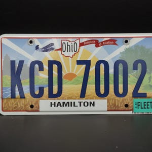 May include: Ohio license plate with the text "KCD 7002" in blue. The plate features a landscape design with a sun, airplane, and the text "Hamilton" at the bottom. The plate is rectangular with a white border.