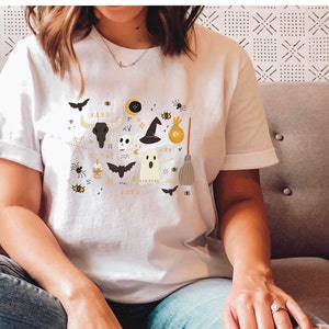 May include: White t-shirt with a Halloween-themed graphic. The design includes bats, a skull, a witch's hat, a ghost, a broom, and the word "LAXD" in gold lettering. The shirt is worn by a person.