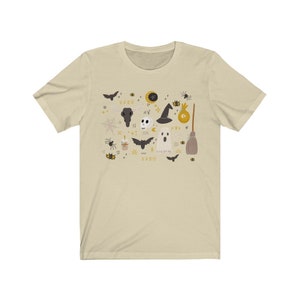 May include: Beige t-shirt featuring a Halloween-themed graphic. The design includes bats, a skull, a witch hat, a ghost, a broom, and other spooky elements in black, gold, and white. The text "1420" is also included in the design.