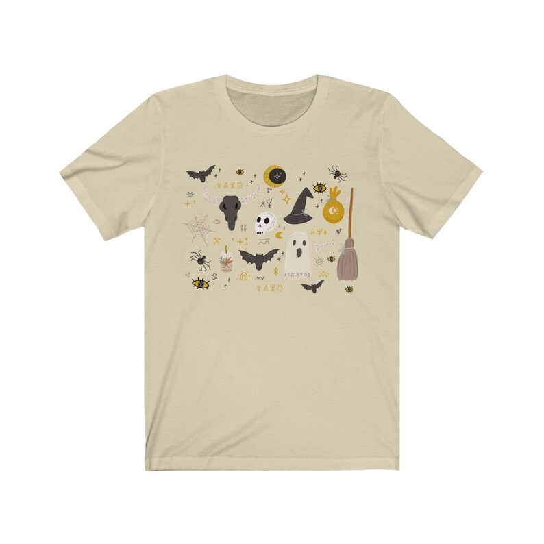 May include: Beige t-shirt featuring a Halloween-themed graphic. The design includes bats, a skull, a witch hat, a ghost, a broom, and other spooky elements in black, gold, and white. The text "1420" is also included in the design.