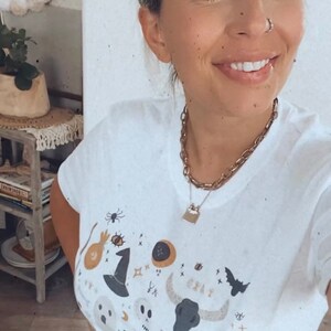 May include: White t-shirt with a Halloween-themed graphic. The design includes a witch's hat, spider, skull, bat, and other spooky elements. A gold chain necklace with a lock pendant is also visible.