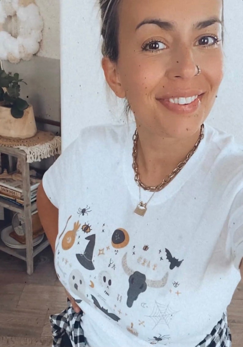 May include: White t-shirt with a Halloween-themed graphic. The design includes a witch's hat, spider, skull, bat, and other spooky elements. A gold chain necklace with a lock pendant is also visible.