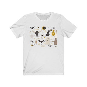 May include: White t-shirt featuring a collection of Halloween-themed illustrations. The design includes a black bull skull, a witch hat, a ghost, bats, spiders, and a broom. The shirt has a crew neck and short sleeves.