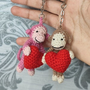 Könnte beinhalten: Zwei gehäkelte Affenschlüsselanhänger, die rote Herzen halten. Die Affen sind rosa und braun und haben lächelnde Gesichter.