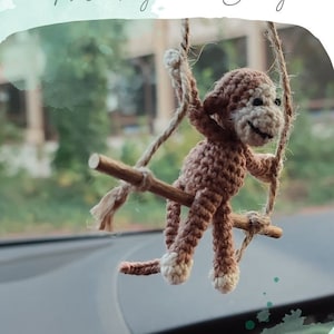 May include: A crocheted monkey toy on a swing, hanging from the rearview mirror of a car. The monkey is brown with a white face and is smiling. The text "Monkey on a Swing" is written above the monkey and the text "@humy_stitch" is written below the monkey.