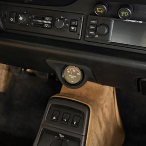 May include: A detailed view of a black car dashboard featuring a radio, air vents, and various controls. A circular gauge is positioned centrally below the dashboard. The interior has a tan color scheme. The car's brand name is visible.