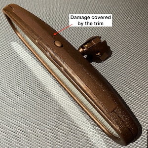 May include: A brown car rearview mirror with a rectangular shape and a small button. The mirror has a metal frame and a mounting bracket. The text "Damage covered by the trim" is visible.
