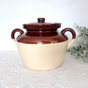 May include: A two-handled ceramic crock with a lid, featuring a glossy dark brown glaze on the upper portion and a smooth cream-colored finish on the lower body. The vessel has a rounded shape with two curved side handles and a matching lid.