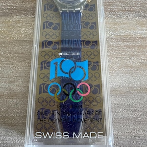 May include: A Swiss-made wristwatch in a clear plastic case. The watch features a dark blue strap with the text "Atlanta, Barcelona, Sarajevo" and Olympic rings. The case reads "1894-1994 ONE HUNDRED YEARS OF THE OLYMPIC MOVEMENT."