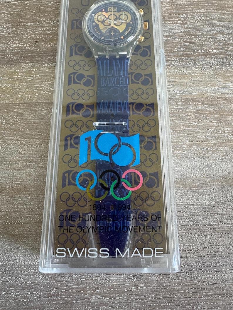 May include: A Swiss-made wristwatch in a clear plastic case. The watch features a dark blue strap with the text "Atlanta, Barcelona, Sarajevo" and Olympic rings. The case reads "1894-1994 ONE HUNDRED YEARS OF THE OLYMPIC MOVEMENT."