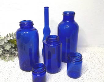 May include: A collection of five cobalt blue glass bottles and jars of varying heights and widths. The set includes two large wide-mouth jars, one tall slender bottle, one medium-sized jar, and one small jar, all featuring a smooth finish.