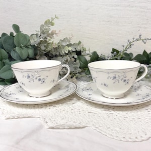 May include: Two white teacups and saucers with a delicate blue floral pattern and silver trim. The cups and saucers are arranged on a white lace tablecloth, with greenery in the background. A classic tea set.