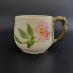 May include: Cream-colored ceramic mug with a pink floral design and green leaves. The handle has a brown, textured appearance. The mug is a rounded shape, suitable for coffee or tea.