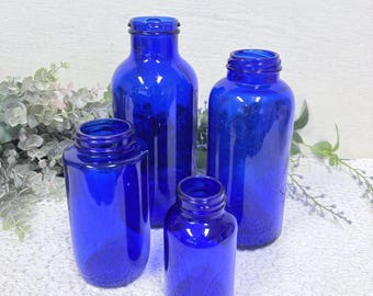 May include: Four cobalt blue glass bottles of varying sizes are arranged on a white lace surface. The bottles have open tops and cylindrical shapes. The bottles are empty and reflect light, showcasing their deep blue color.