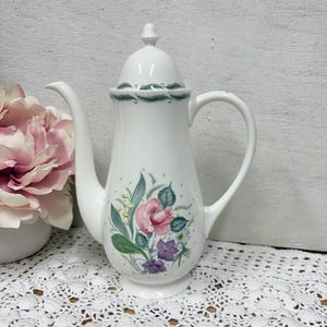 May include: White ceramic coffee pot with a floral design featuring a pink rose, purple flowers, and green leaves. The pot has a curved spout, a handle, and a lid with a small finial. The rim of the lid has a green scalloped design.
